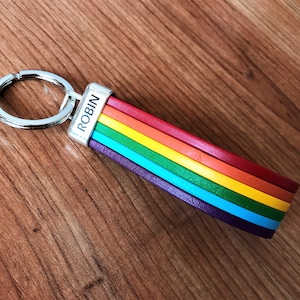 Könnte beinhalten: Ein regenbogenfarbener Leder-Schlüsselanhänger mit einem silbernen Ring und dem Namen "ROBIN" auf dem Metallanhänger.