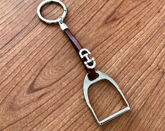 May include: A silver stirrup-shaped keyring with a brown leather strap and a silver ring. The stirrup has a small silver detail in the shape of a horseshoe.