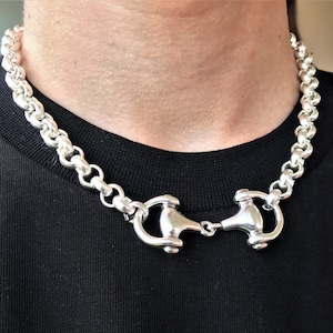May include: A silver chain necklace with a large, chunky curb chain design. The necklace has a unique, double-looped clasp.