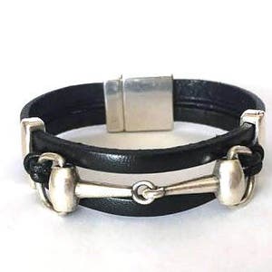 Pode incluir: Uma pulseira de couro preto com um fecho magnético e um charme de freio de cavalo prateado.