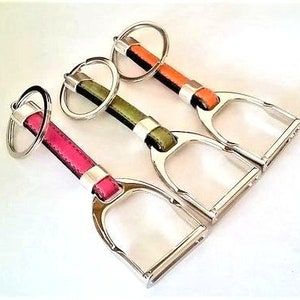 Puede incluir: Tres llaveros en forma de estribo con correas de cuero. Los llaveros son de color rosa, verde y naranja.