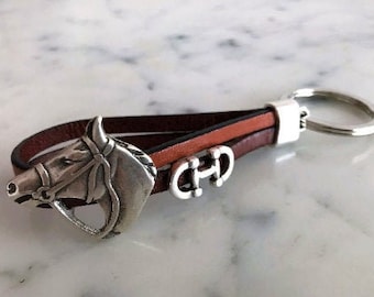 May include: A brown leather key chain with a silver horse head charm and a silver key ring. The horse head charm has a bridle and a bit.