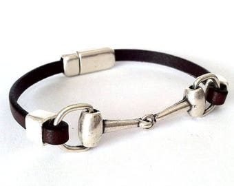 Pode incluir: Um bracelete de couro castanho com um fecho de prata em forma de freio de cavalo. O bracelete tem um fecho magnético.