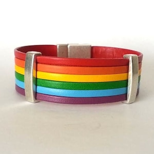 May include: A rainbow leather bracelet with a silver magnetic clasp. The bracelet is made of six different coloured leather strips: red, orange, yellow, green, blue, and purple.