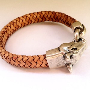May include: A braided leather bracelet with a silver animal head clasp. The bracelet is light brown and the clasp is detailed with a textured surface.