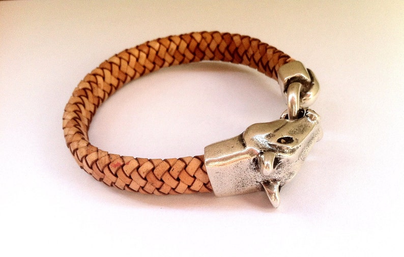 May include: A braided leather bracelet with a silver animal head clasp. The bracelet is light brown and the clasp is detailed with a textured surface.