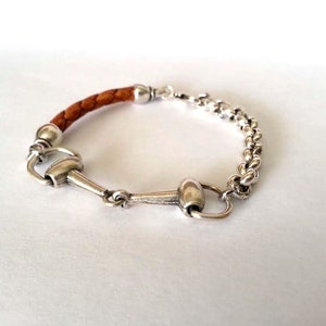Könnte beinhalten: Ein braunes Lederarmband mit einer silbernen Kette und einem silbernen Pferdebiss-Anhänger.