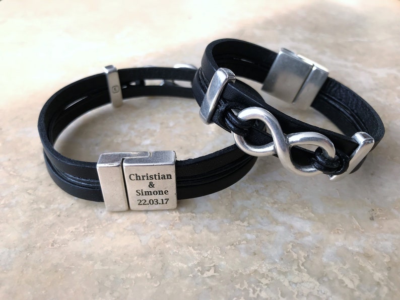 May include: Two black leather bracelets with silver infinity symbol charms and a silver clasp. One bracelet has the text "Christian & Simone 22.03.17" engraved on the clasp.