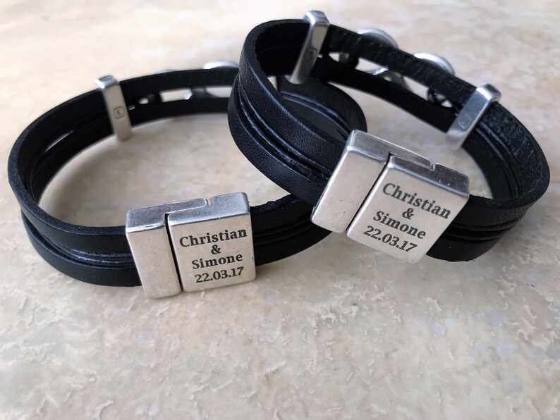 May include: Two black leather bracelets with silver clasps. Each clasp has the text "Christian & Simone 22.03.17" engraved on it.