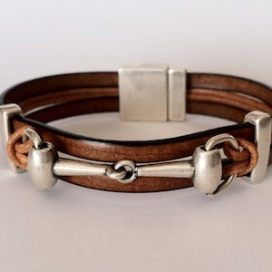 May include: A brown leather bracelet with a silver equestrian bit charm. The bracelet has a magnetic clasp.