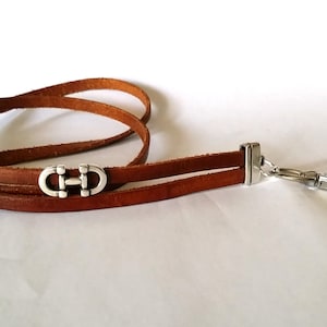 May include: A brown leather lanyard with two silver metal clasps and a silver metal hook. The clasps are shaped like the letter "H" with a circle around it.