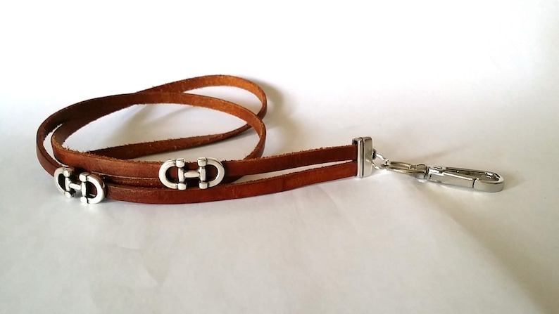 May include: A brown leather lanyard with two silver metal clasps and a silver metal hook. The clasps are shaped like the letter "H" with a circle around it.