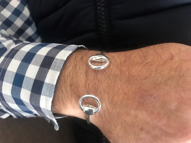 May include: A silver bracelet with two circular, open-ended charms. The bracelet is made of a black leather strap.
