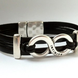 Può includere: Braccialetto in pelle nera con un ciondolo in argento a forma di simbolo di infinito. Il ciondolo ha la parola "INFINITY" incisa.