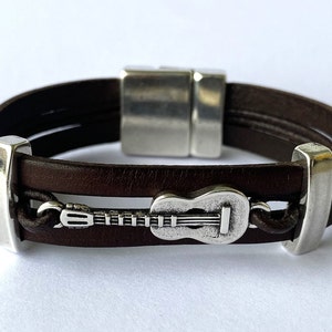 Könnte beinhalten: Ein braunes Lederarmband mit einem silbernen Gitarrenanhänger und einem silbernen Magnetverschluss.