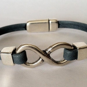 K&ouml;nnte beinhalten: Ein graues Lederarmband mit einem silbernen Unendlichkeits-Anh&auml;nger und einem Magnetverschluss.