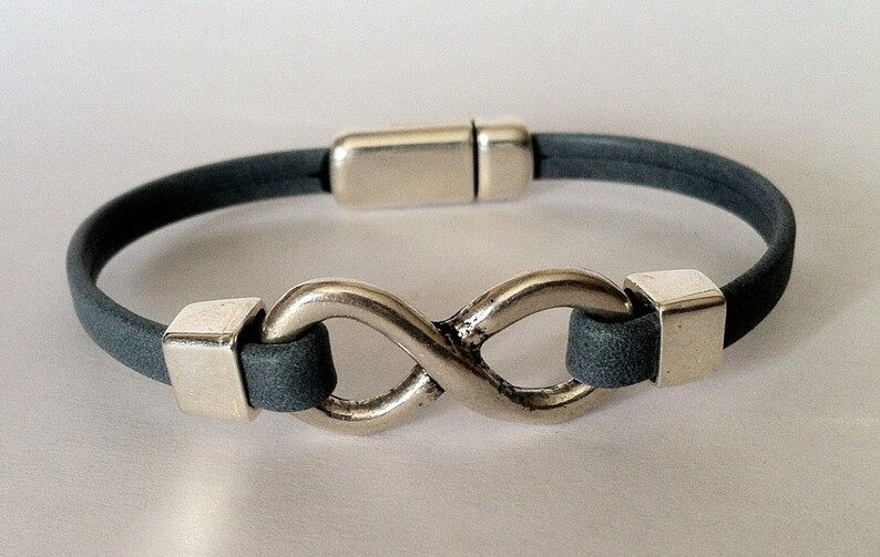 K&ouml;nnte beinhalten: Ein graues Lederarmband mit einem silbernen Unendlichkeits-Anh&auml;nger und einem Magnetverschluss.