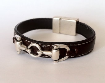 Pode incluir: Um bracelete de couro castanho com um fecho magnético prateado e um design de freio de cavalo prateado.
