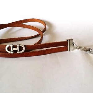 May include: A brown leather lanyard with a silver clasp and three silver decorative charms. The charms are shaped like a double 'C' with a horizontal bar connecting the two 'C's.