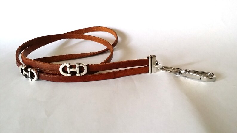 May include: A brown leather lanyard with a silver clasp and three silver decorative charms. The charms are shaped like a double 'C' with a horizontal bar connecting the two 'C's.