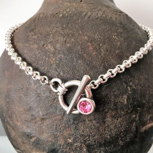 May include: A silver-toned necklace with a toggle clasp and a pink gemstone. The necklace features a chain of small, rounded links. The toggle clasp has a circular loop and a bar closure. The gemstone is a round, bright pink color.