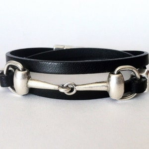Könnte beinhalten: Ein schwarzes Lederarmband mit einem silbernen Reitsporn-Detail. Das Armband ist doppelt gewickelt und hat ein schlichtes, elegantes Design.