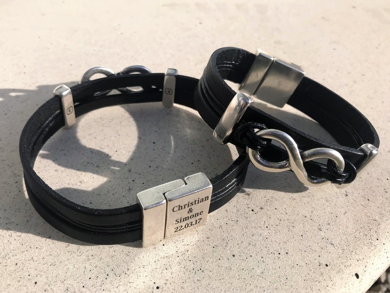 May include: Two black leather bracelets with silver infinity symbol charms. One bracelet has a silver clasp engraved with "Christian & Simone 22.03.17".