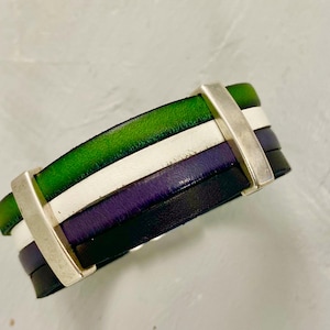May include: A multi-colored leather bracelet with green, white, purple, and black bands. The bracelet is held together by two silver-colored rectangular bars. The leather appears to be high-quality and the bracelet is likely handmade.