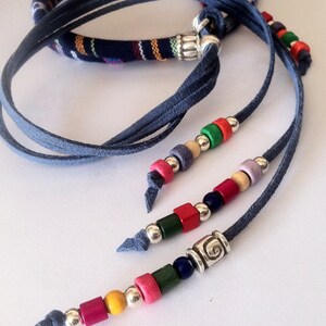 Puede incluir: Un collar de m&uacute;ltiples hebras con cordones de gamuza azul y cuentas de madera en varios colores, incluyendo rojo, verde, azul, amarillo y morado. El collar presenta un dije de plata con un dise&ntilde;o de espiral.