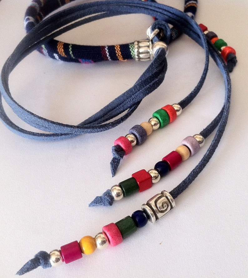 Puede incluir: Un collar de m&uacute;ltiples hebras con cordones de gamuza azul y cuentas de madera en varios colores, incluyendo rojo, verde, azul, amarillo y morado. El collar presenta un dije de plata con un dise&ntilde;o de espiral.