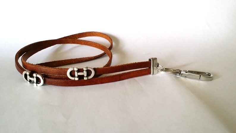 May include: A brown leather lanyard with two silver metal charms in the shape of the letter "H". The lanyard has a silver metal clip on one end.