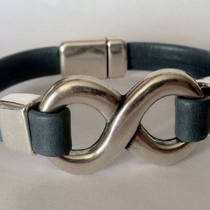 K&ouml;nnte beinhalten: Ein graues Lederarmband mit einem silbernen Unendlichkeits-Symbol als Verschluss.