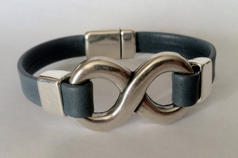 K&ouml;nnte beinhalten: Ein graues Lederarmband mit einem silbernen Unendlichkeits-Symbol als Verschluss.
