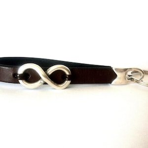 Puede incluir: Un llavero de cuero marrón con un dije de plata en forma de símbolo de infinito y un anillo de llave de plata.