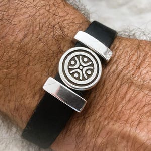 Może przedstawiać: Bransoletka ze skóry w kolorze czarnym z srebrnym zawieszką przedstawiającą stylizowany wzór czterech osób trzymających się za ręce. Bransoletka ma dwa srebrne metalowe akcenty.