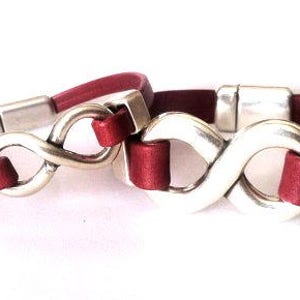 May include: Two red leather bracelets with silver infinity symbol charms. The charms are connected by a silver chain.