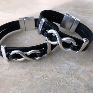 May include: Two black leather bracelets with silver infinity symbol charms. The bracelets have magnetic clasps.