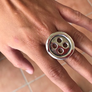 May include: A silver ring with a large button-shaped design featuring five circular cutouts. The ring is worn on a person's finger.