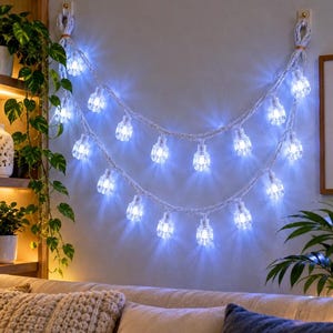 May include: A decorative string of lights featuring two draped strands of thick white rope with integrated clear, faceted bulb covers that emit a bright, cool-toned white glow. The rope is secured to the wall at two points with small hooks.