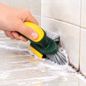 Puede incluir: Un cepillo de limpieza manual con mango ergonómico amarillo, base verde oscuro y cerdas grises rígidas. El cepillo cuenta con una pequeña cuchilla de goma amarilla en el lateral, utilizada para fregar juntas con jabón en superficies de azulejos.