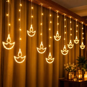 Puede incluir: Una cortina de luces LED de color blanco cálido con siluetas de lámparas diya colgantes. Las luces están dispuestas en hilos verticales suspendidos de un cable horizontal, y cada hilo termina en un contorno de diya brillante y flameante.
