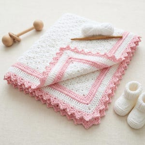 Op de afbeelding: Een wit gehaakt dekentje met een roze rand en geschulpte boorden, vergezeld door een paar witte gebreide babyslofjes, een houten rammelaar, een bol witte wol en een houten haaknaald, allemaal gerangschikt op een neutraal oppervlak.