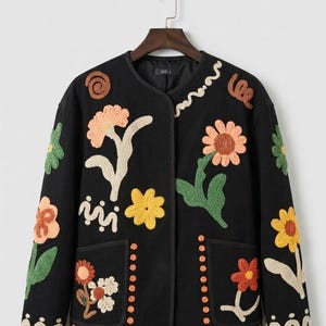 May include: A black long-sleeved jacket featuring a crew neckline and two front pockets. The garment is decorated with colorful, textured floral embroidery in shades of orange, yellow, and green, along with small orange circular accents.