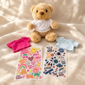 Könnte beinhalten: Ein beigefarbener Plüschteddybär in einem weißen T-Shirt, dazu ein pinkes T-Shirt, ein hellblaues T-Shirt und zwei Aufkleberbögen mit bunten Motiven, darunter Texte wie "GOOD VIBES", "BE KIND", "OUT OF THIS WORLD" und "EXPLORE".