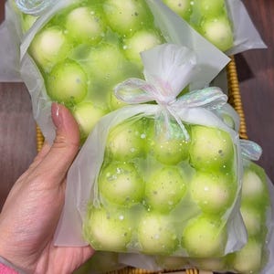 Puede incluir: Una vela cuadrada de color verde claro con textura de burbujas, empaquetada en una bolsa de plástico transparente. La bolsa está atada en la parte superior con una cinta iridiscente translúcida. La vela presenta una cuadrícula de pequeñas esferas redondeadas con un acabado brillante.
