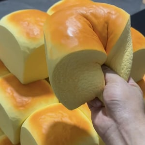以下が含まれることがあります： 黄金色の焼き色がついた皮と淡い黄色の内側を持つ、四角い形の柔らかいフォーム製パンのコレクション。一つのパンが握られており、その柔軟で弾力のある質感と、圧縮されて元の形に戻る様子が示されています。