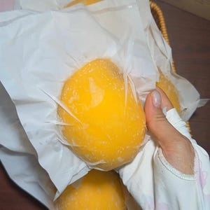 Puede incluir: Varias frutas redondas de color amarillo brillante están envueltas individualmente en papel blanco fino y translúcido. Las frutas tienen una piel suave y vibrante y están dispuestas juntas en una cesta tejida, mostrando su forma uniforme.
