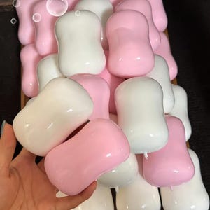 Könnte beinhalten: Eine Ansammlung von glatten, abgerundeten, sanduhrförmigen Riegeln in den Farben Pink und Weiß. Diese Objekte haben eine glänzende Oberfläche und einen kleinen, mittigen Vorsprung an einem Ende. Die Riegel sind in einem dichten Haufen angeordnet.