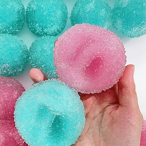 Könnte beinhalten: Eine Ansammlung runder, texturierter Kugeln, die mit funkelnden Zuckerkristallen überzogen sind. Die Kugeln sind hauptsächlich in leuchtendem Türkis und kräftigem Pink gehalten und weisen in der Mitte eine kleine kreisförmige Vertiefung auf.