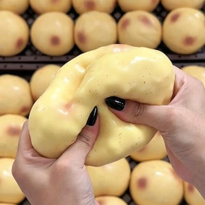 Pode incluir: Um brinquedo macio, de cor amarelo-pálido, em forma de batata com pequenas manchas castanhas. A superfície é lisa e flexível, permitindo que seja comprimida e deformada sob pressão. Faz parte de uma coleção de brinquedos idênticos.